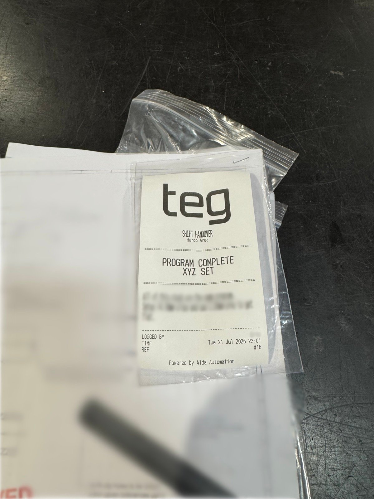 A printed handover tag attached to a bag of machined parts on a machine bed, resting on the job drawing. The drawing and the body of the note are blurred.